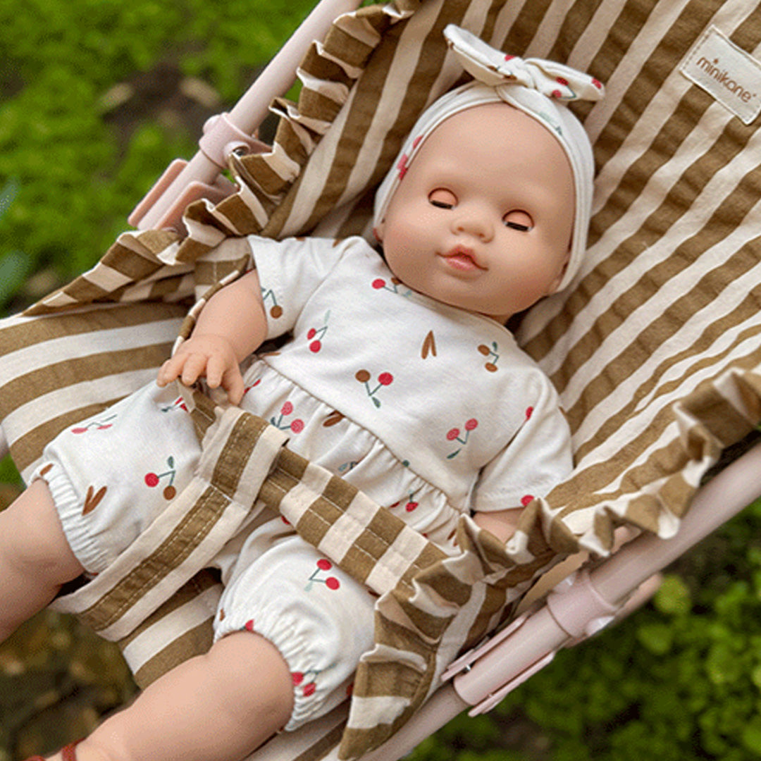 minikane babypop slaapt in een gestreepte kinderwagen, gekleed in een wit pakje met kersenprint.
