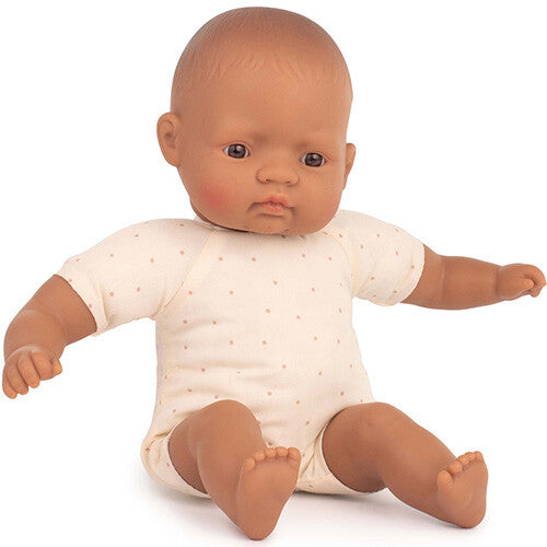 Miniland babypop met bruine huidskleur en stippenrompertje zit op de grond.
