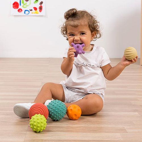 Een baby sabbelt op een paarse miniland sensorische bal en houdt een beige bal vast, met andere ballen op de vloer.