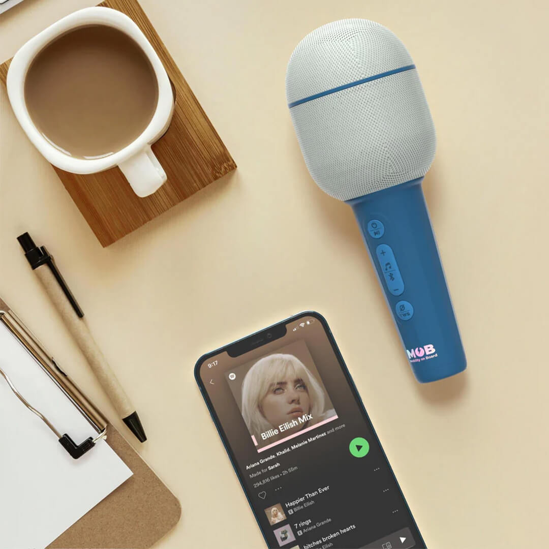 Mob karaoke microfoon naast een smartphone met muziekapp, een koffiekop en pen op een bureau.