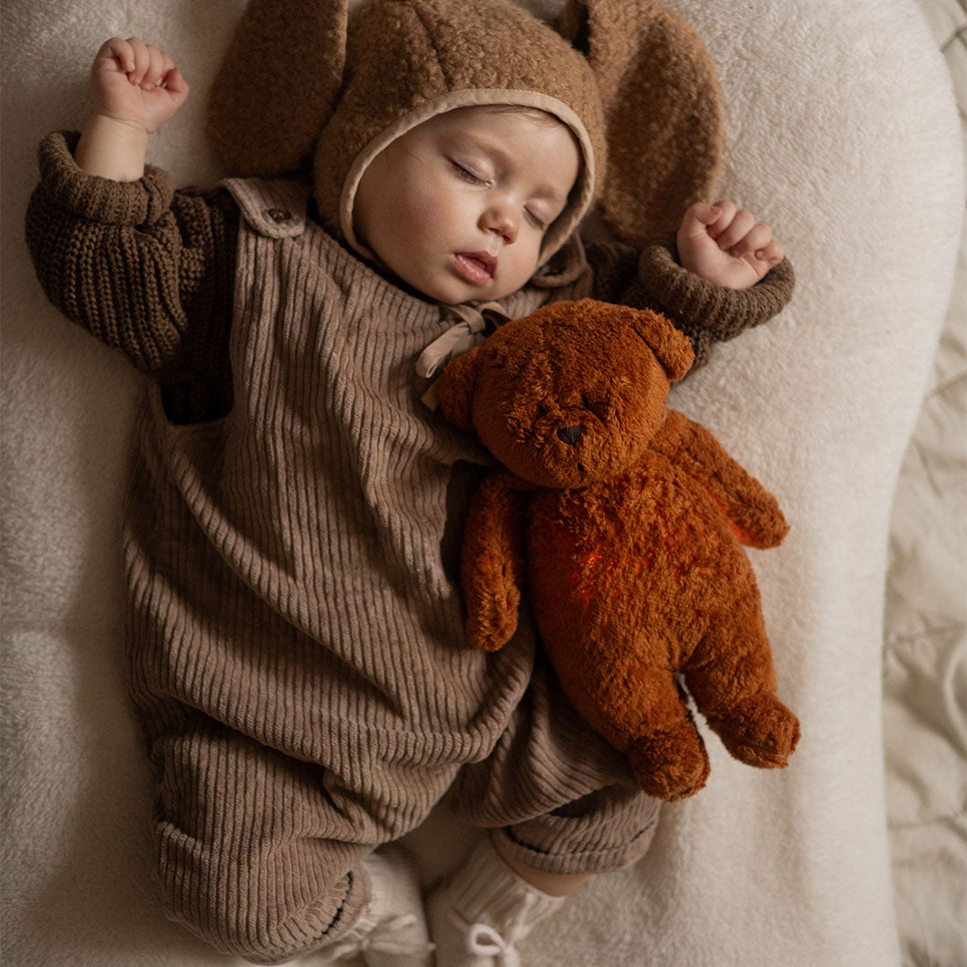 Een slapende baby in een bruin pakje knuffelt de Moonie knuffelbeer met oranje gloed op de buik.