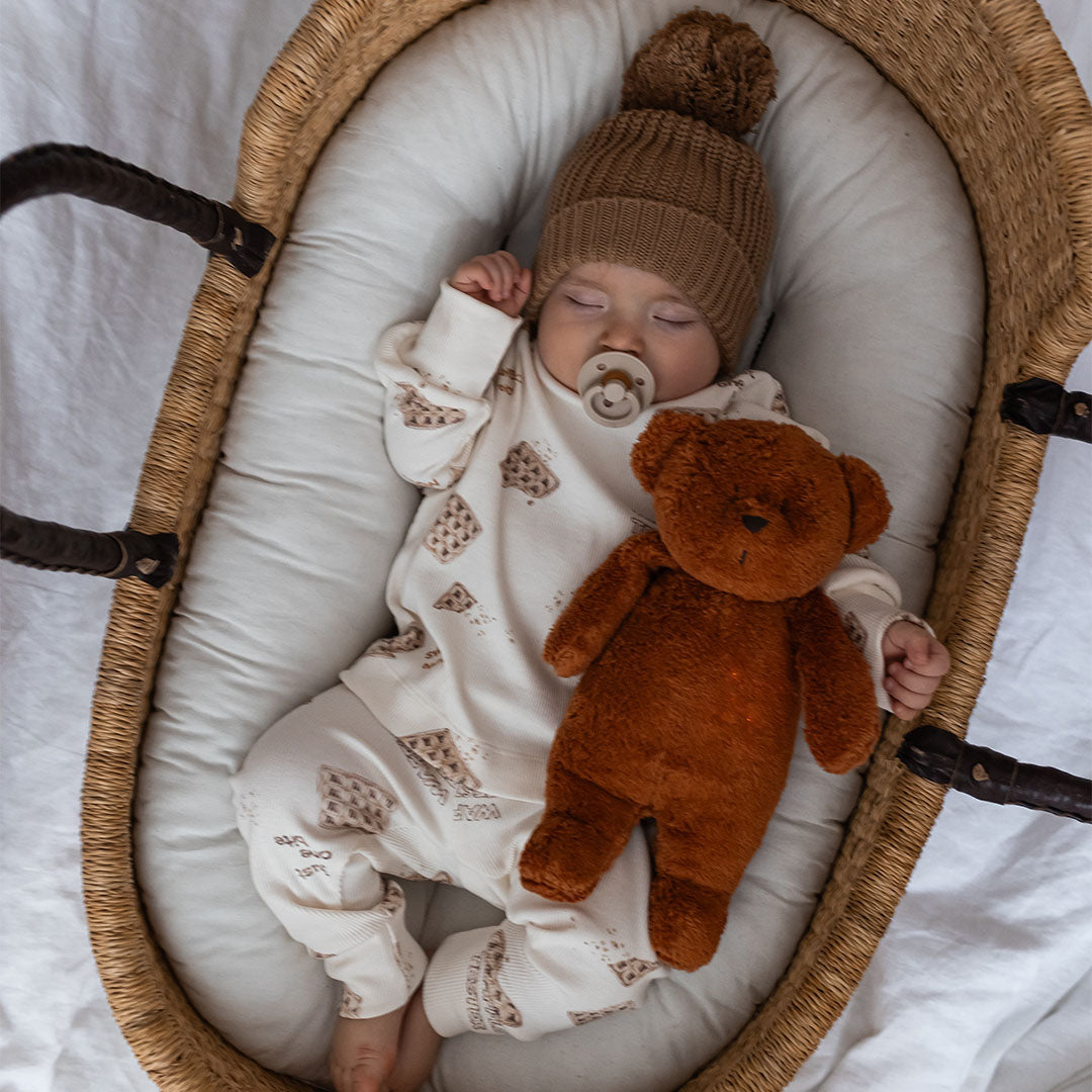 Een slapende baby met speen ligt in een wiegje naast de Moonie knuffelbeer met oranje gloed op de buik.