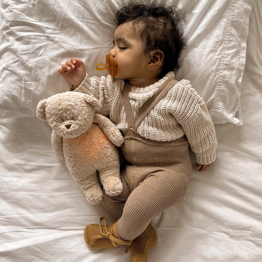 Slapende baby met een Moonie pluchen knuffelbeer met oranje gloed op een wit bed.