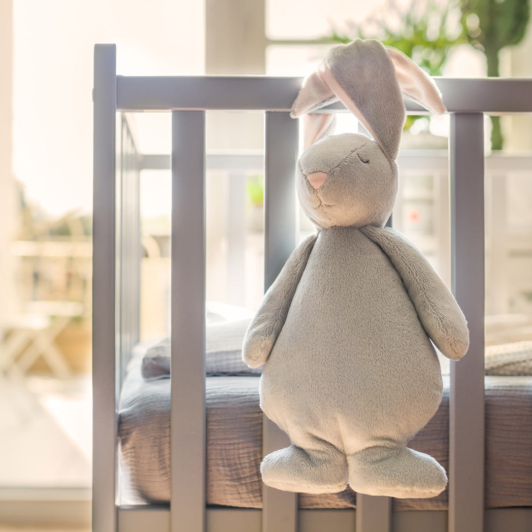Het moonie pluchen konijn hangt over de rand van een grijze babywieg.