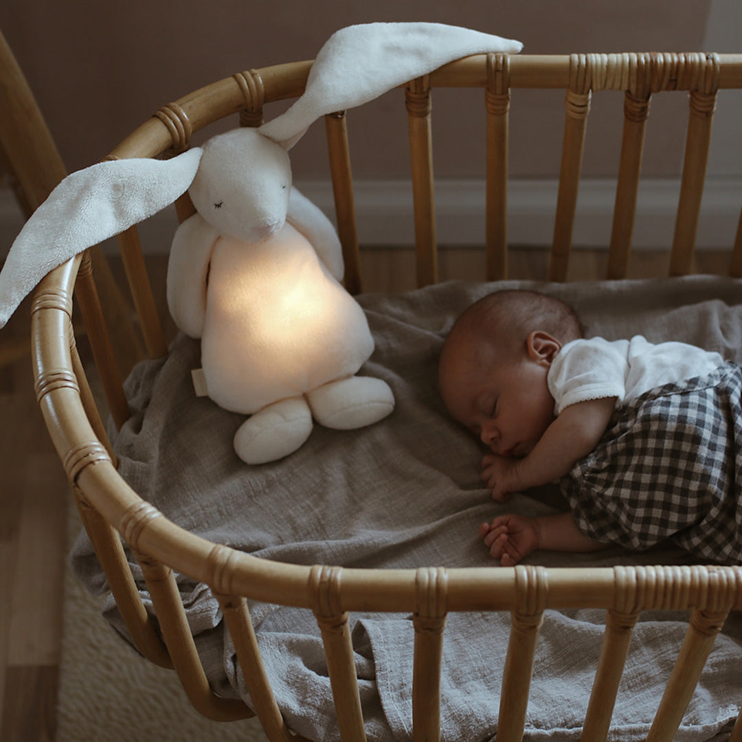 Een slapende baby ligt in een wieg naast het moonie knuffelkonijn met nachtlamp, dat zachtjes gloeit.