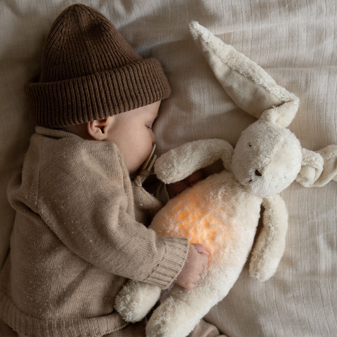 Een slapende baby knuffelt het Moonie pluchen konijn met oplichtende buik.