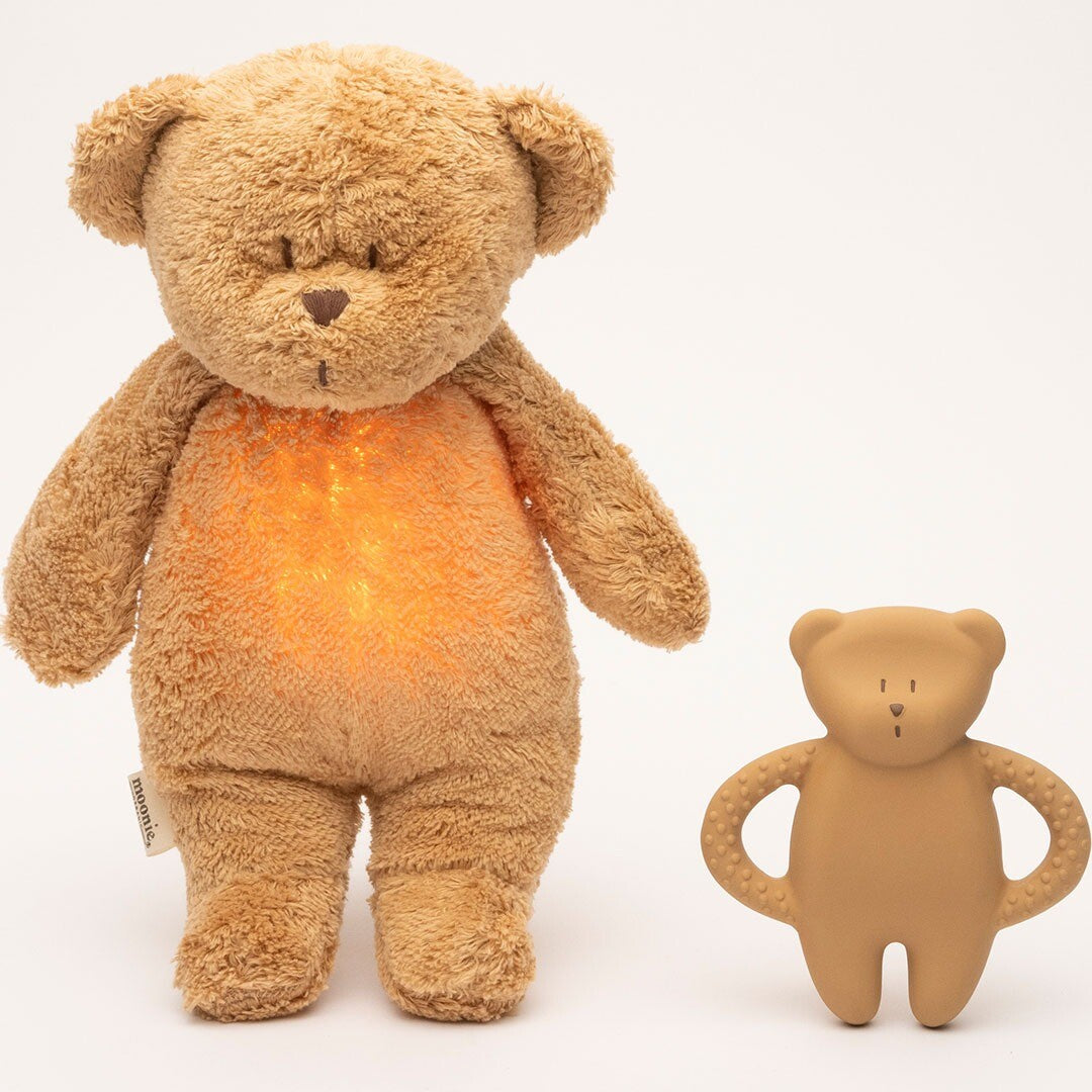 Een moonie rubberen bijtring in de vorm van een beige beer staat naast een grotere, lichtgevende knuffelbeer.