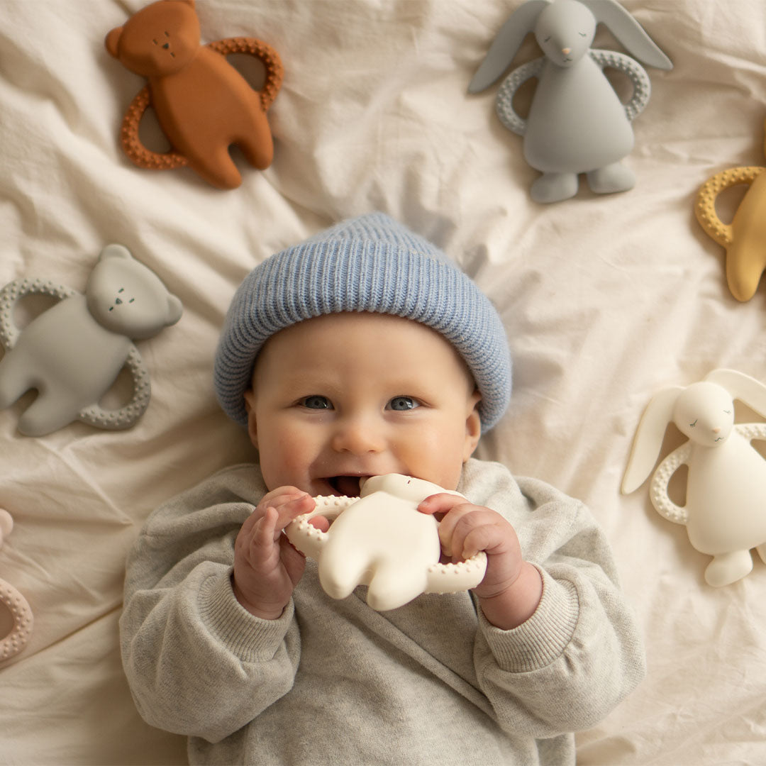 Een lachende baby met blauwe muts kauwt op een moonie konijnen bijtring, omringd door andere bijtringen in de vorm van dieren.