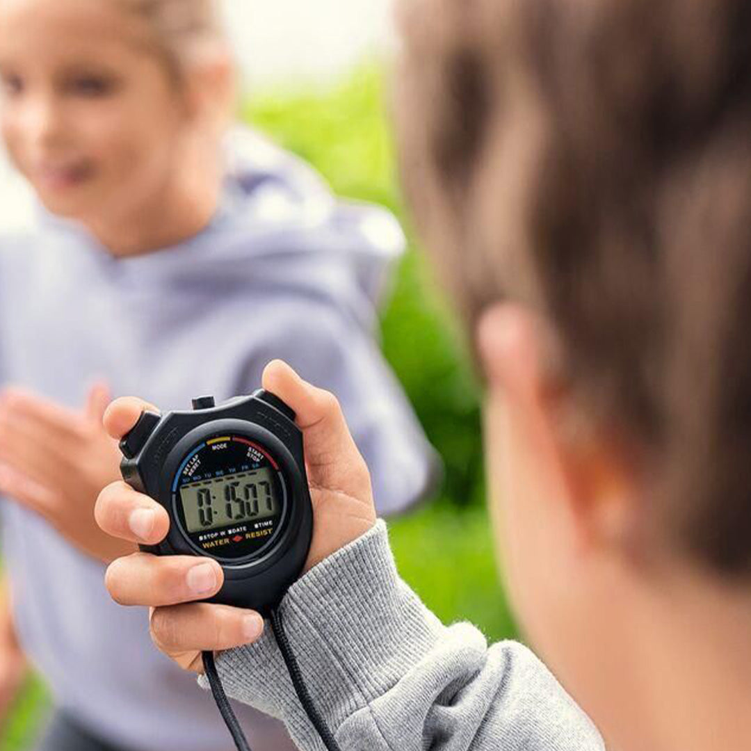 Een hand houdt de zwarte digitale stopwatch vast, terwijl op de achtergrond een persoon rent.
