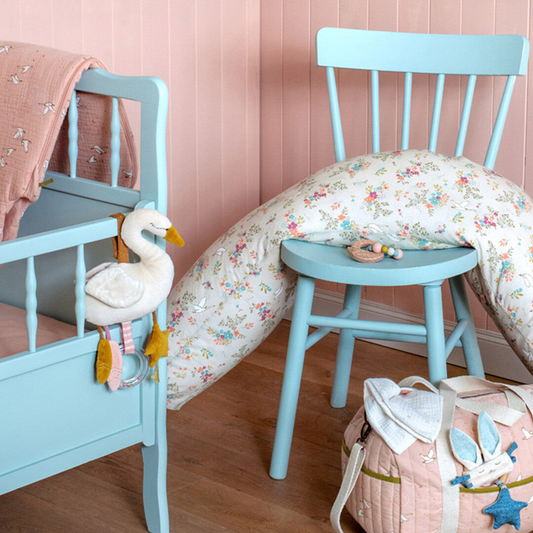De zwaan activiteitenknuffel hangt aan een babywieg in een kinderkamer met een blauwe stoel en roze tas.