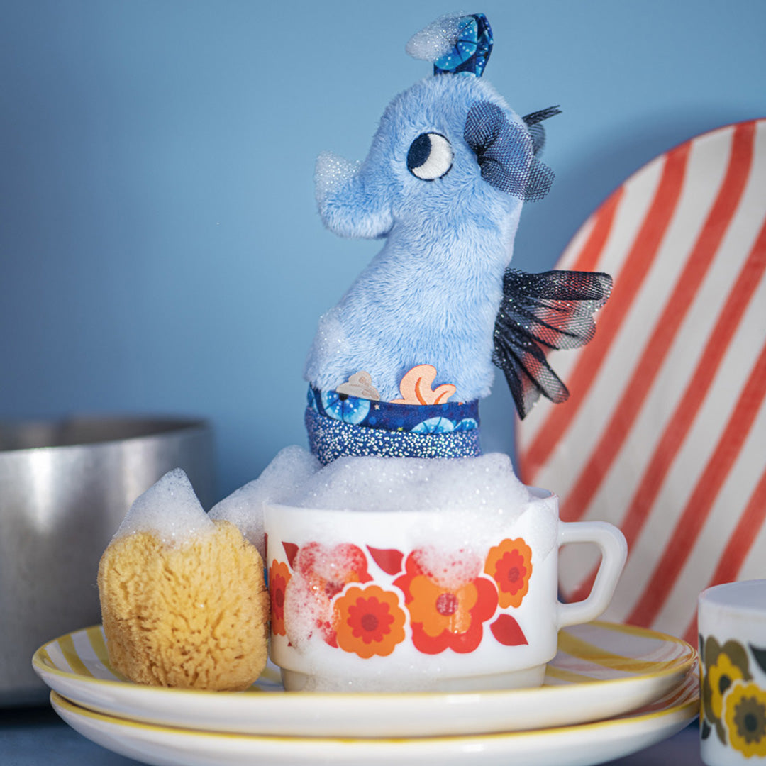 Blauw pluche zeepaardje activiteitenknuffel staat in een schuimende kop op een stapel borden.