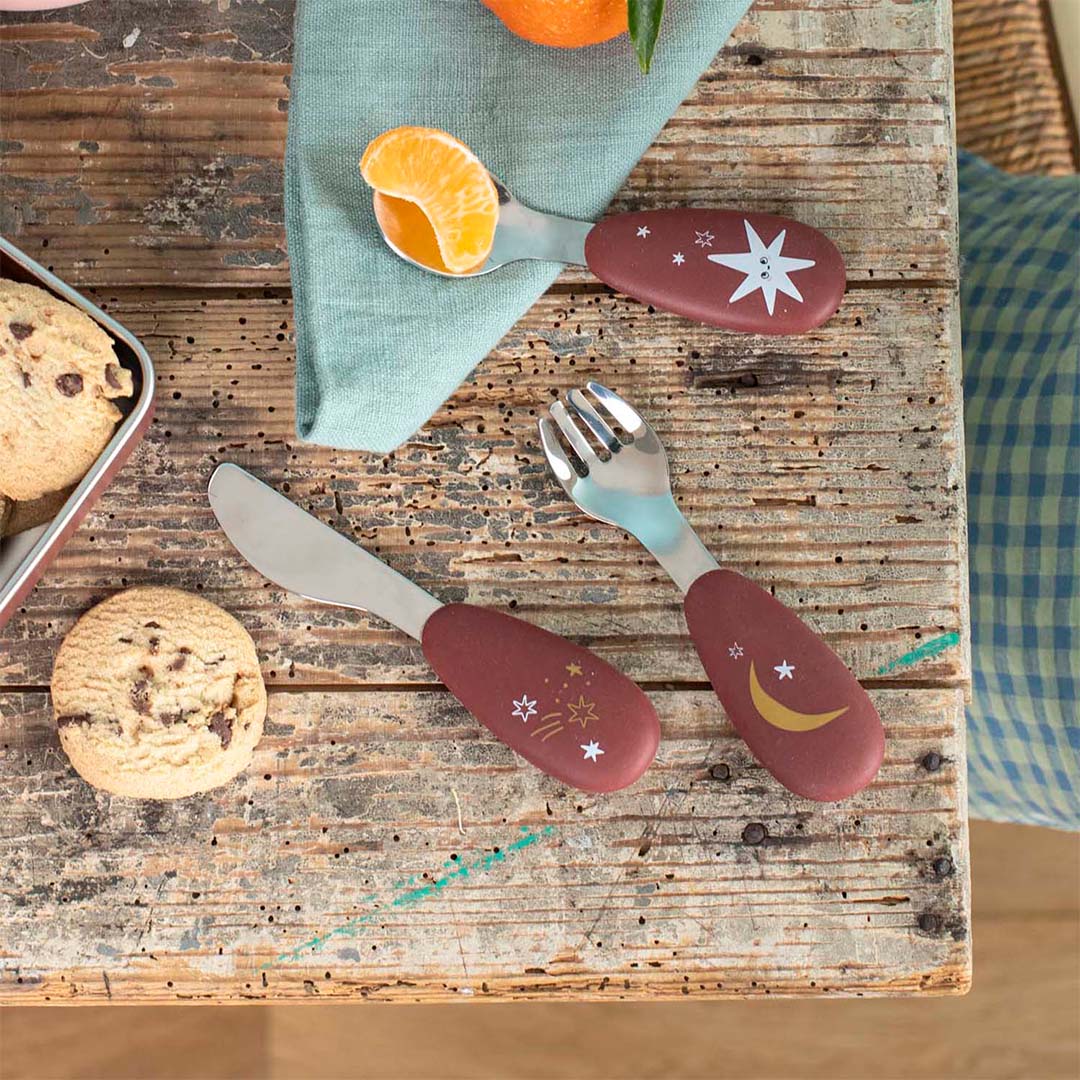 Moulin Roty bestekset met sterren- en maanpatronen op een houten tafel met koekjes en mandarijn.