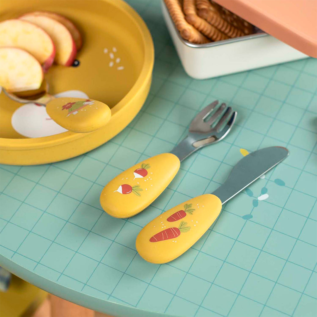 De gele kinderbestekset met radijsjes en wortels ligt op een blauwe tafel naast een gele kom met appel.