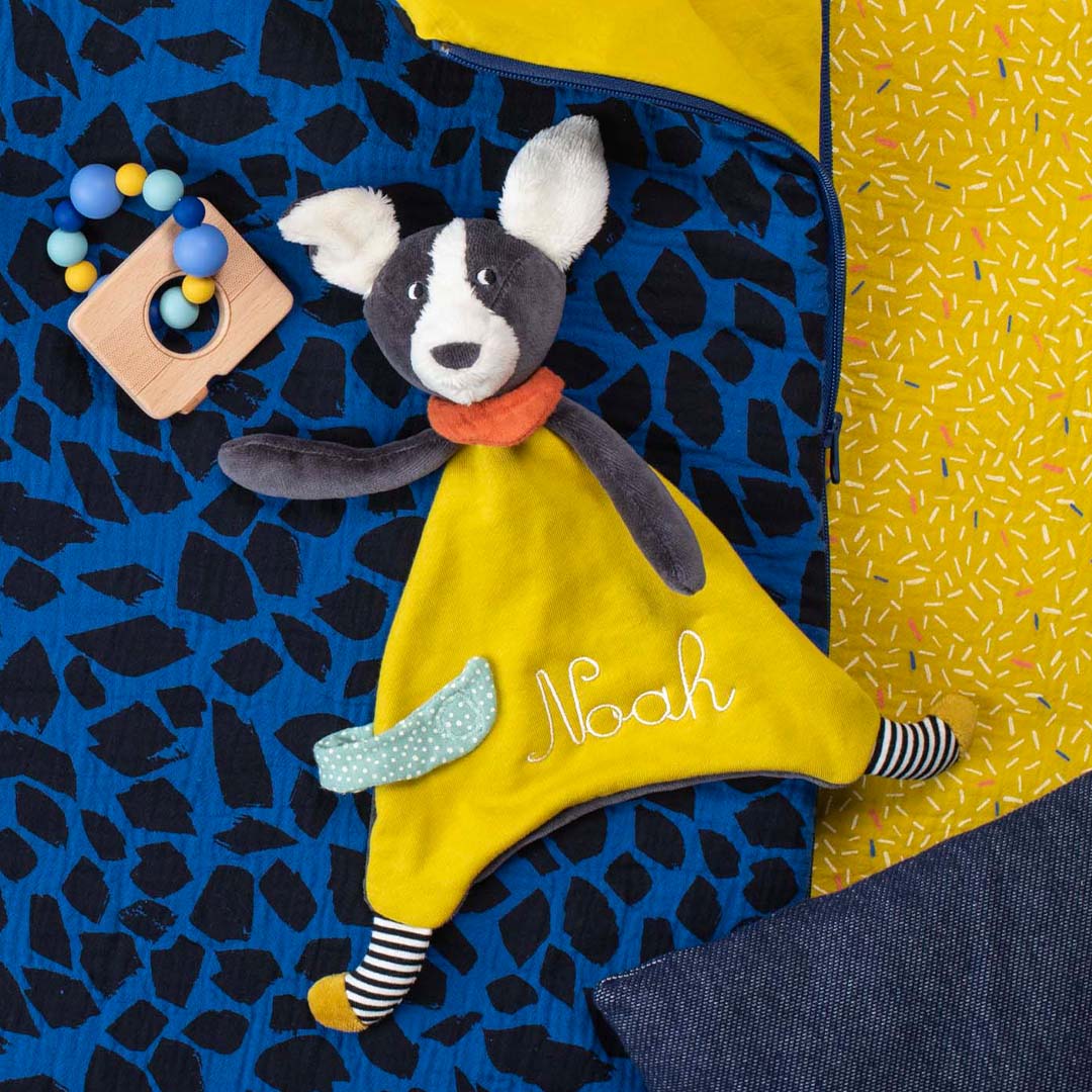 Een Moulin Roty knuffeldoekje met muis en een houten camera bijtring liggen op een blauw-geel patroon.