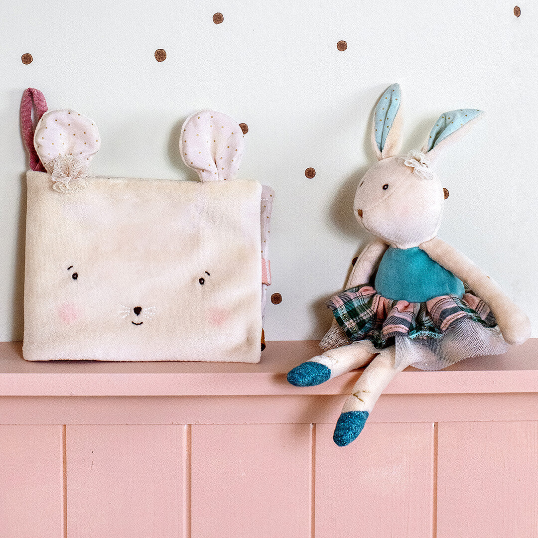 Het beige muis baby fotoalbum staat op een roze plank naast een knuffelkonijn.