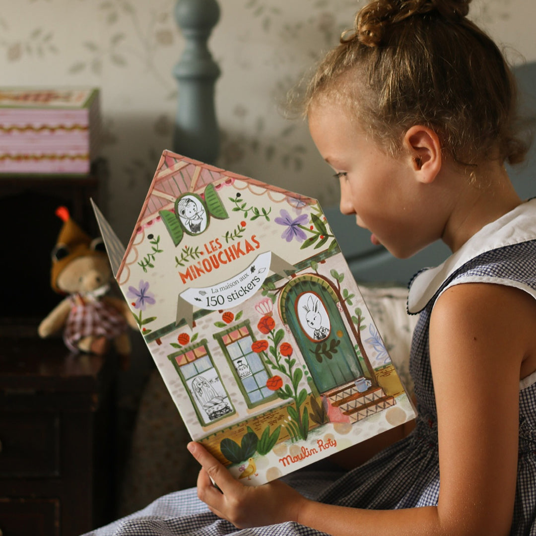 Een meisje bekijkt het Moulin Roty kleur- en stickerboek Les Minouchkas, dat de vorm heeft van een huis.