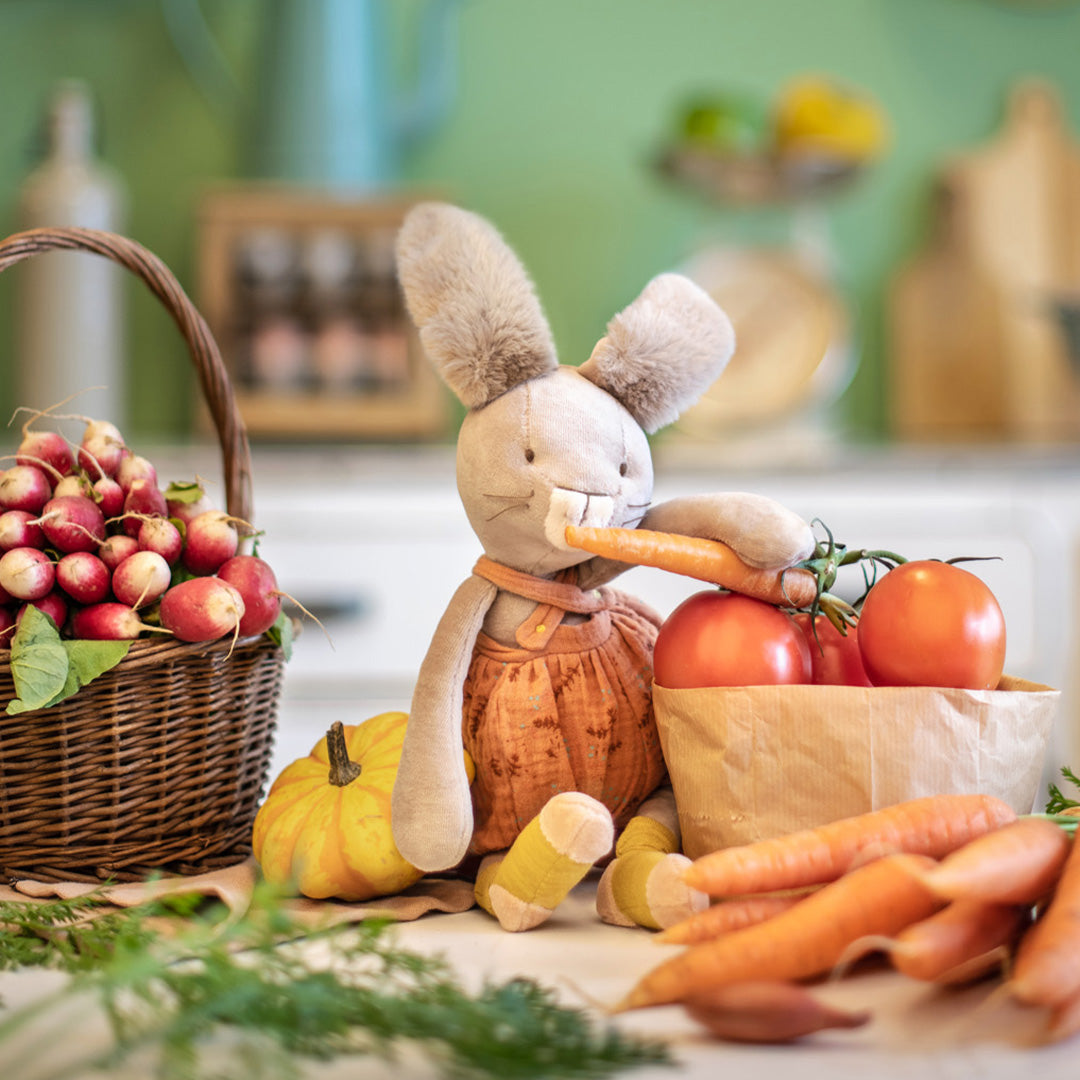 Moulin Roty knuffelkonijn zit tussen groenten en eet een wortel.