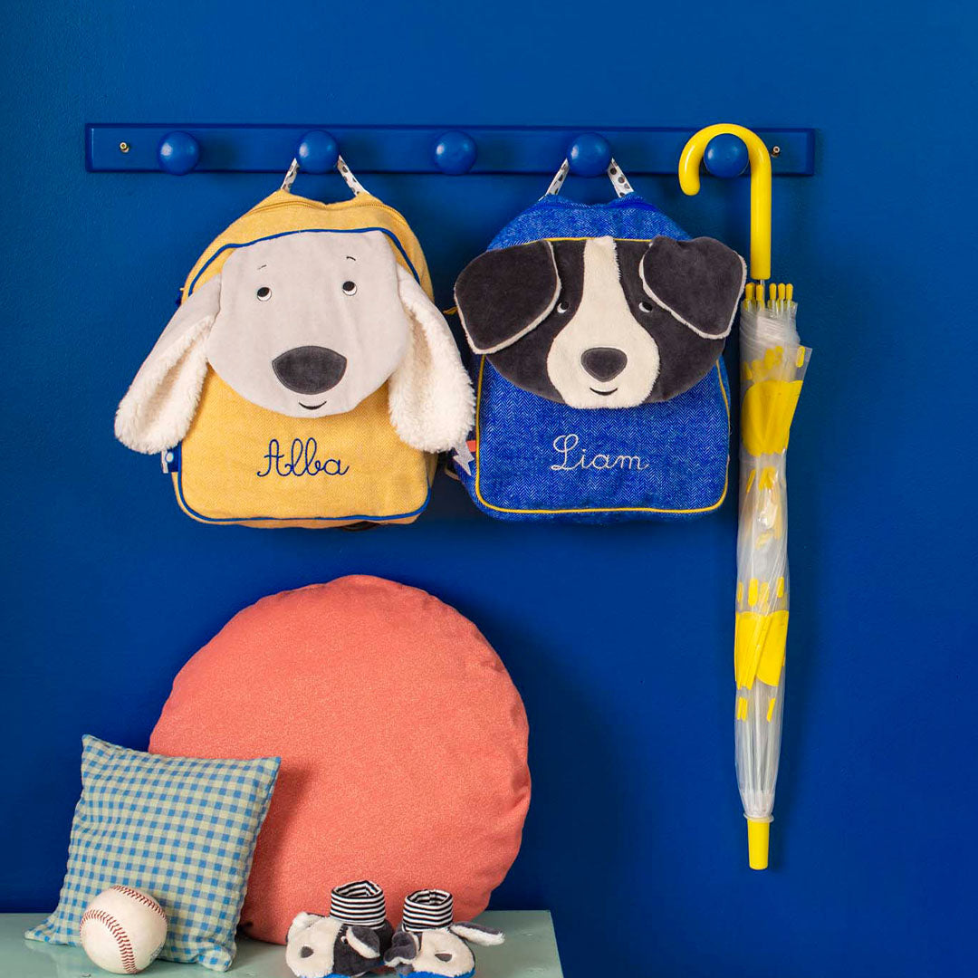 Twee Moulin Roty peuterrugzakken met hondenfiguren, Pilou en Puce, hangen aan een blauwe kapstok naast een gele paraplu.