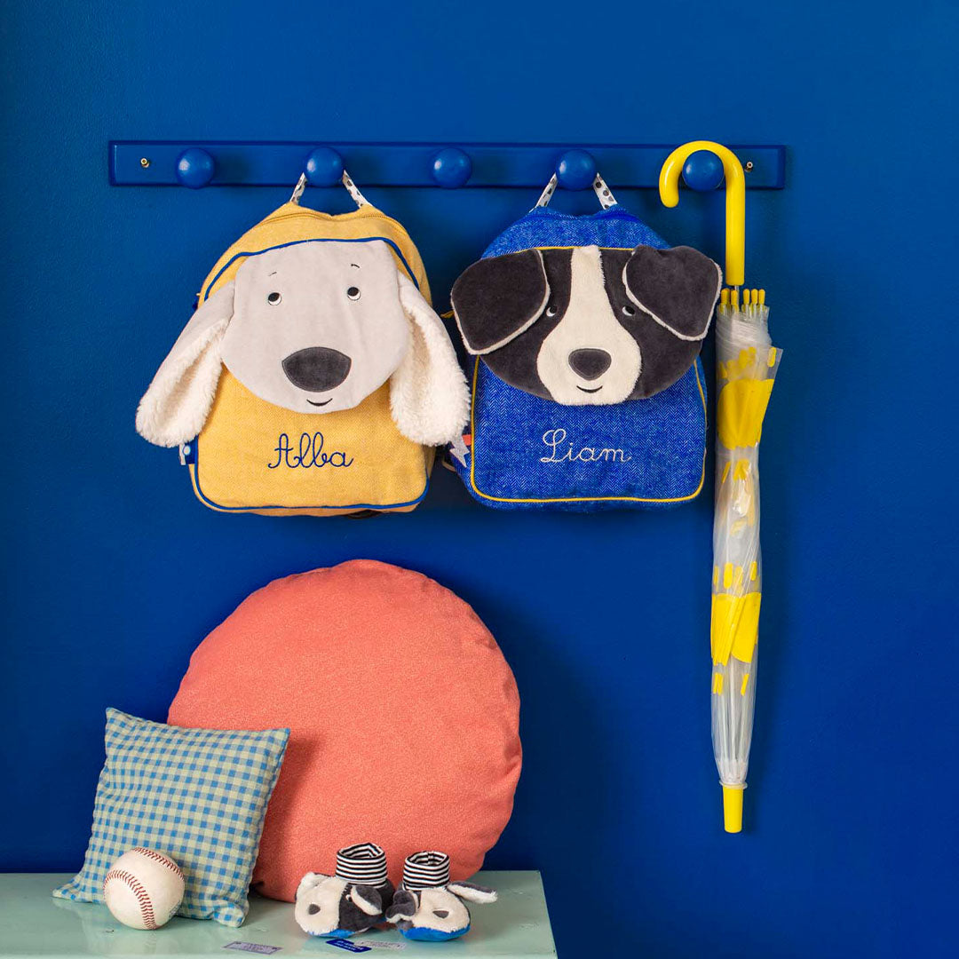 Twee Moulin Roty peuterrugzakken met hondenhoofden, een gele en een blauwe, hangen aan een kapstok naast een gele paraplu.