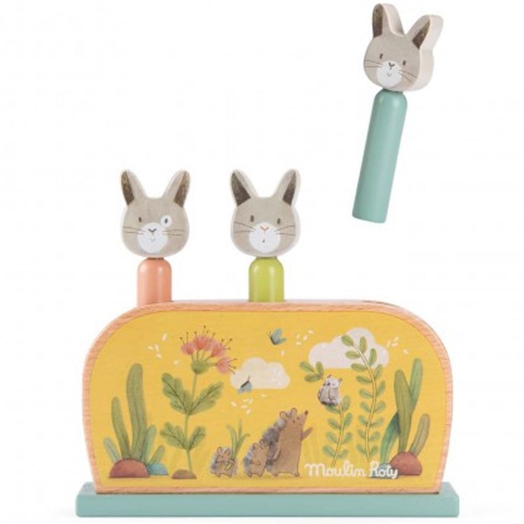 Moulin Roty houten pop-up spel voor baby's met twee konijnen in de gaten en één konijn erboven, op een gele basis met bosdieren en planten.