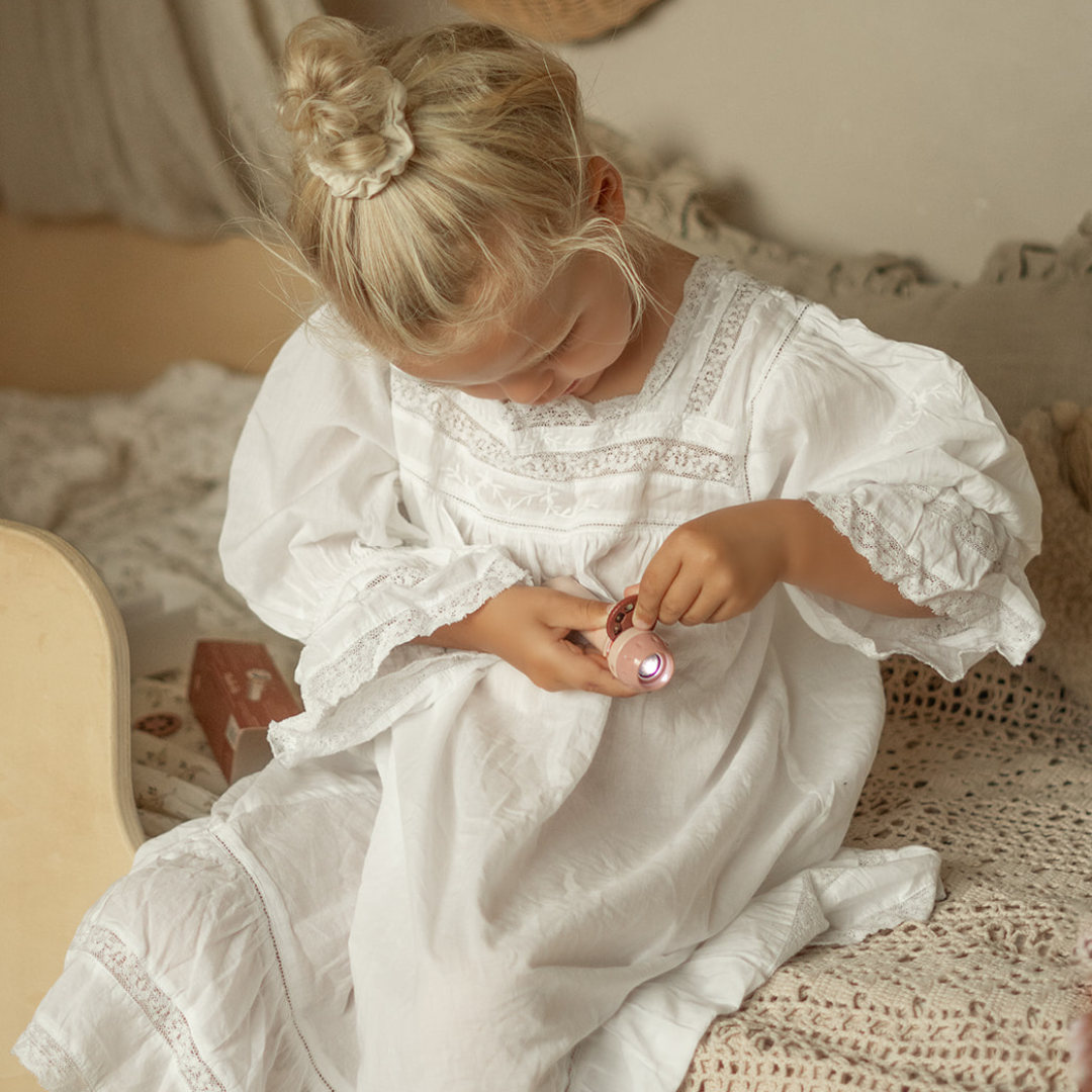 Een meisje in een witte jurk bekijkt de roze Moulin Roty projector in haar handen.