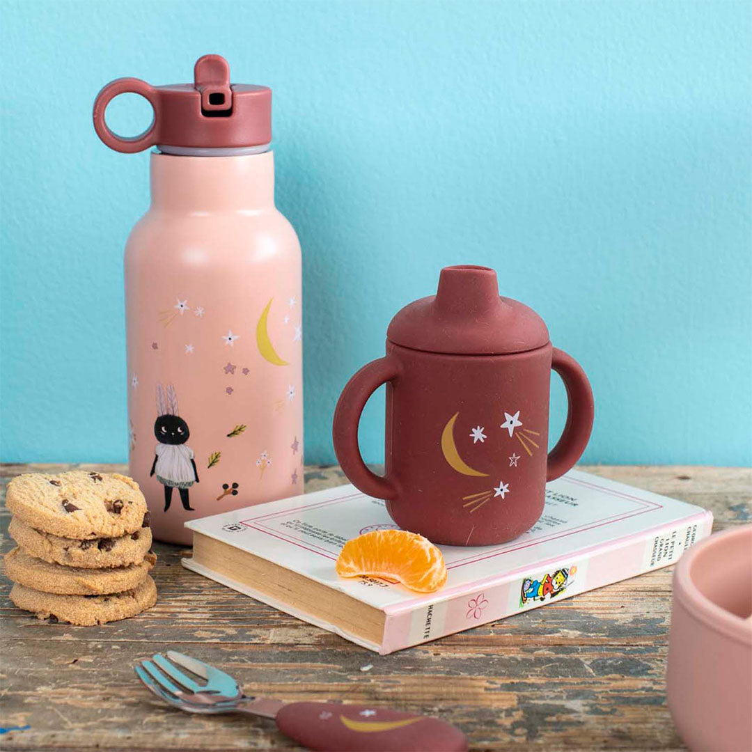 Moulin Roty roze kinderdrinkfles met een figuur en sterren, naast een donkerrode drinkbeker en koekjes op een houten tafel.
