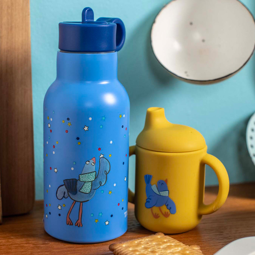Moulin Roty blauwe kinder waterfles met vogelillustratie naast een gele drinkbeker op een houten ondergrond.