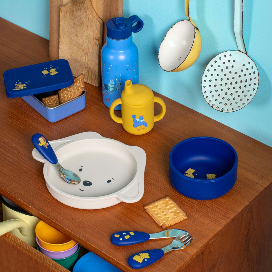 Collectie Moulin Roty kinder eetgerei met blauwe waterfles, lunchbox, gele drinkbeker, bord en bestek op een houten tafel.