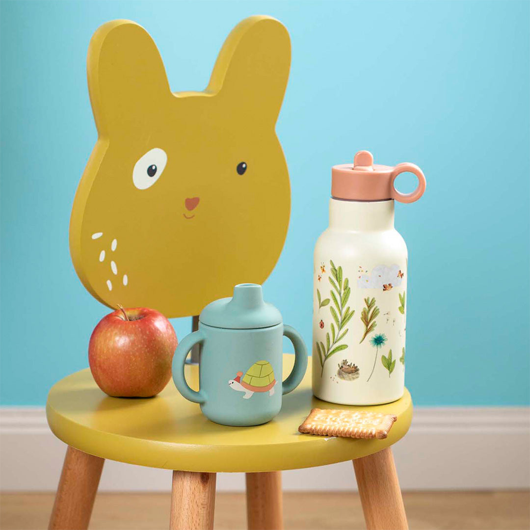 Moulin Roty RVS drinkfles met bladeren- en dierenpatroon staat op een gele kinderstoel naast een blauwe tuitbeker en een appel.
