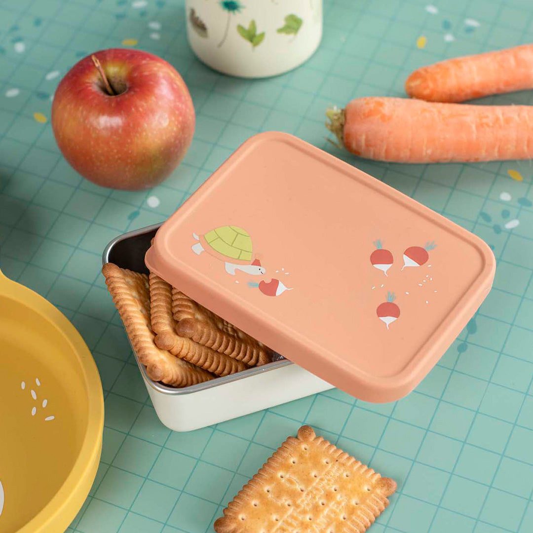 Moulin Roty rvs snackdoosje met oranje deksel, gevuld met koekjes, naast een appel en wortels.