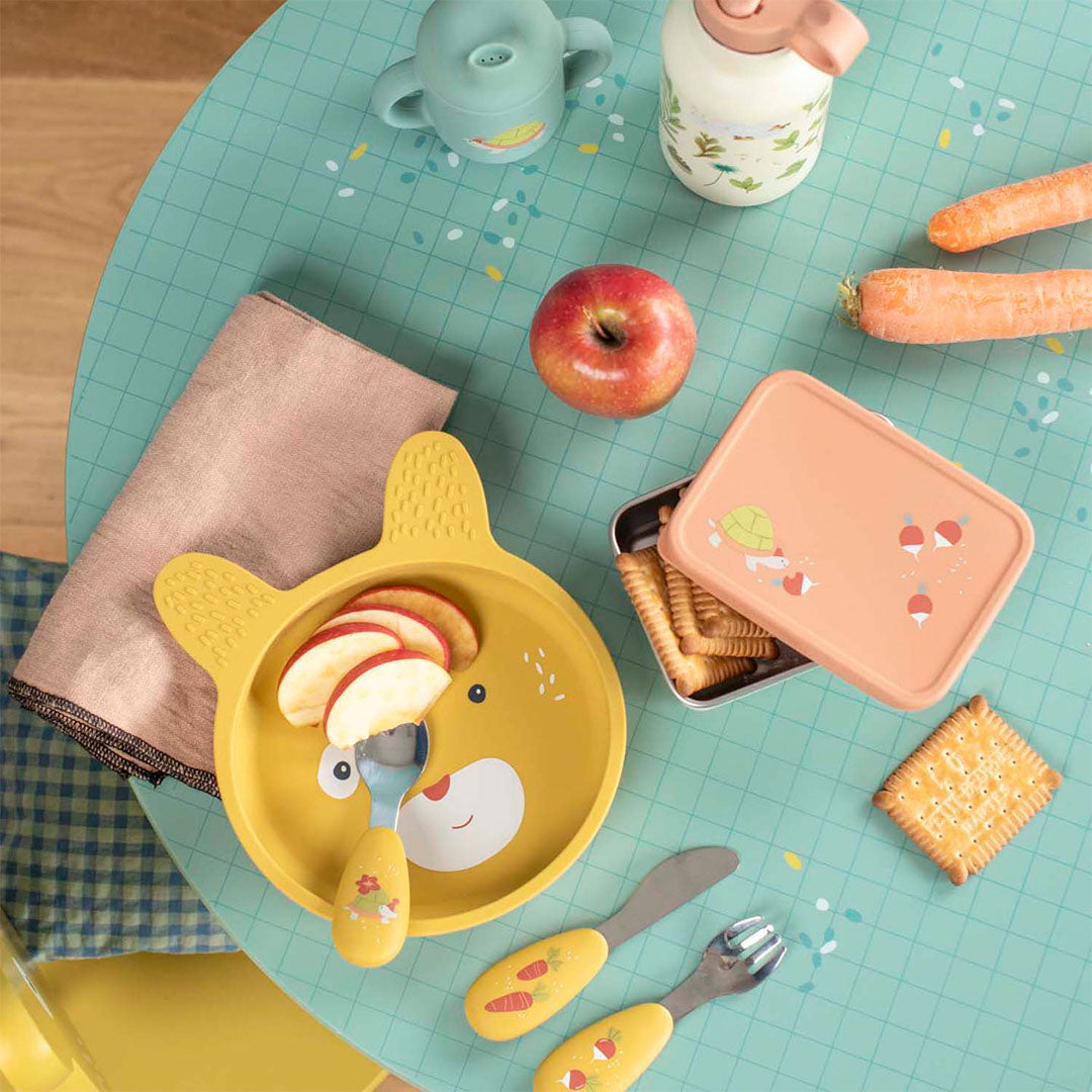 Overzicht van een kindertafel met het Moulin Roty rvs snackdoosje, een geel bord met bestek, een appel en wortels.
