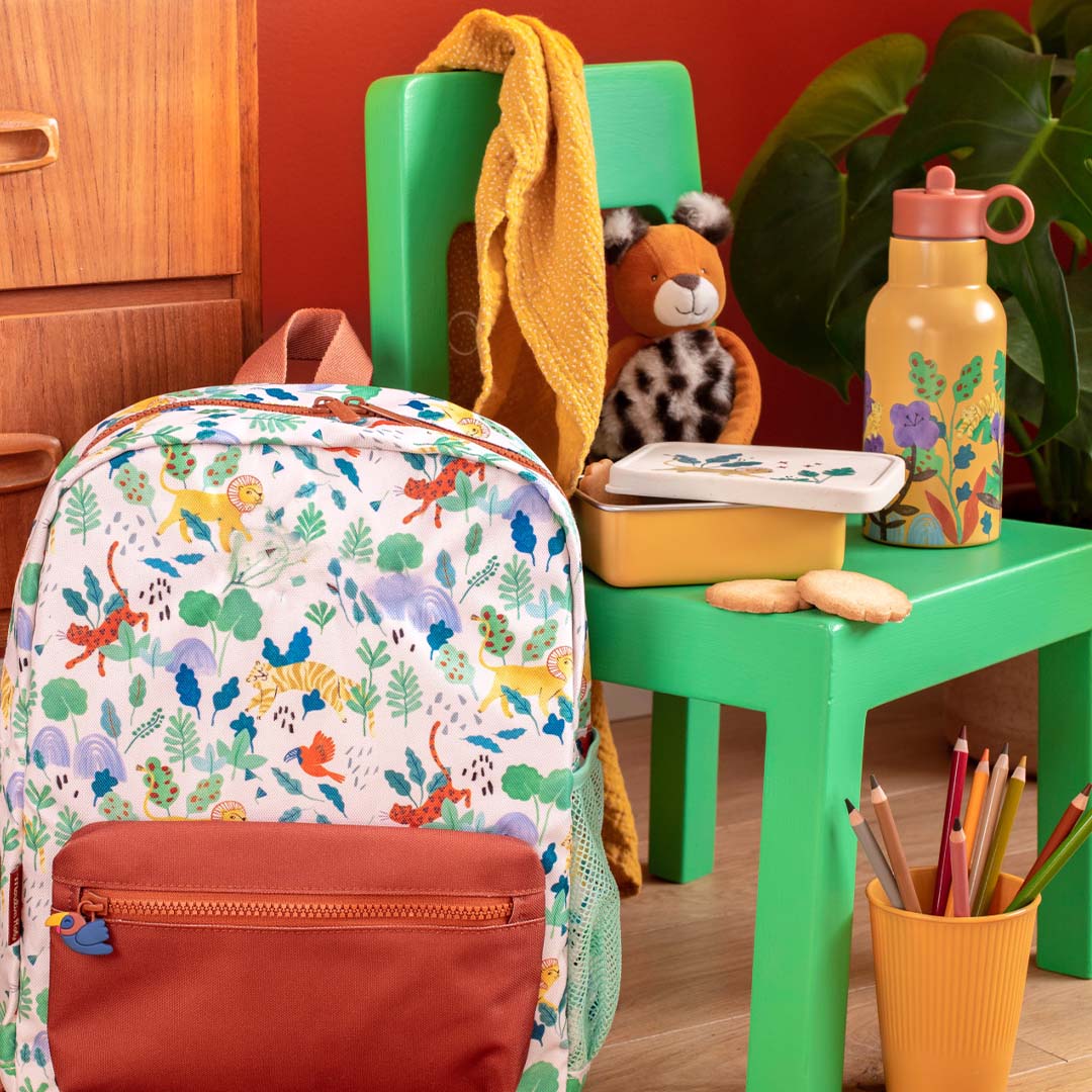 Moulin Roty gele drinkfles op een groen krukje, naast een bijpassende rugzak en lunchbox.