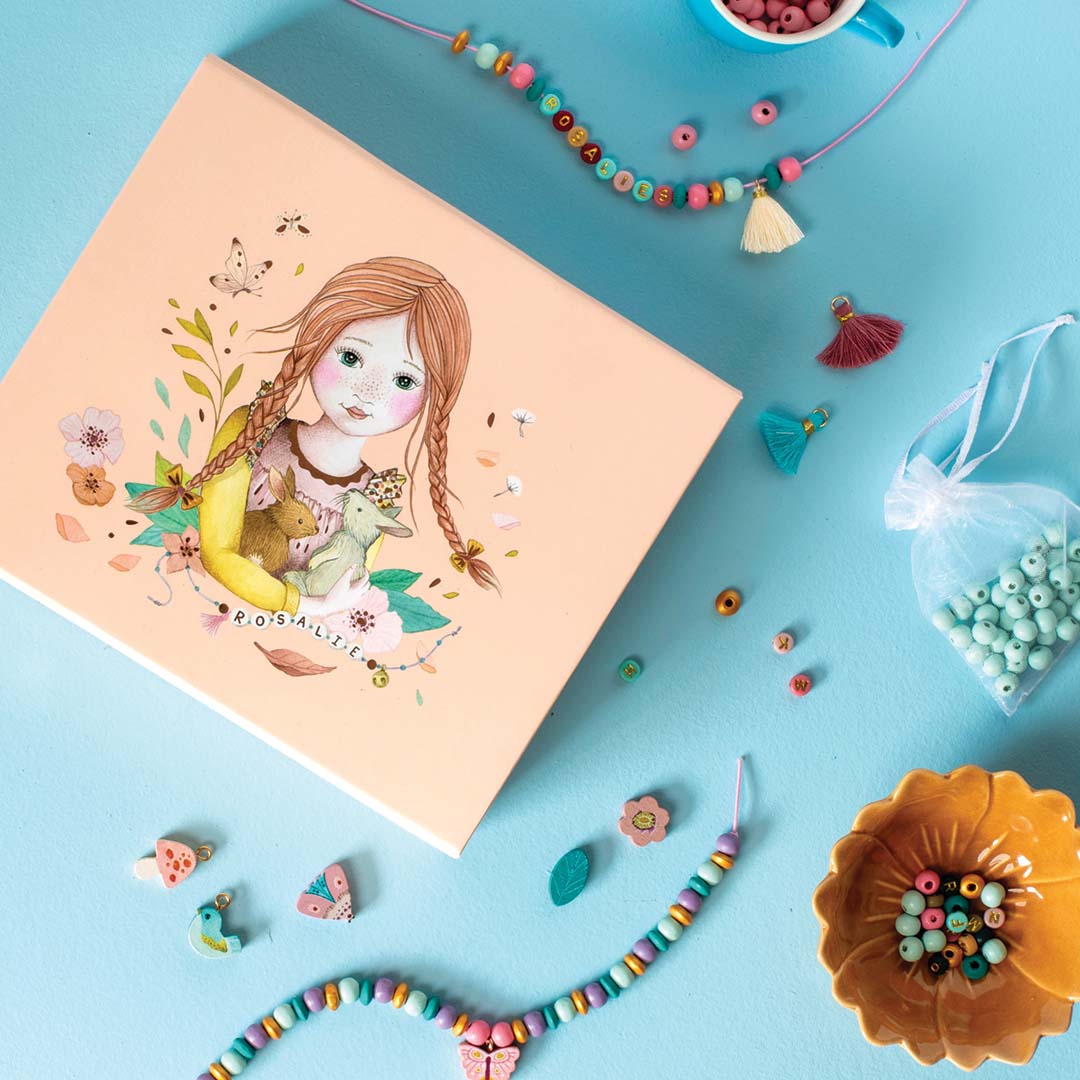 Gesloten verpakking van de Moulin Roty kralenset op een blauwe ondergrond, omringd door kralen en sieraden.