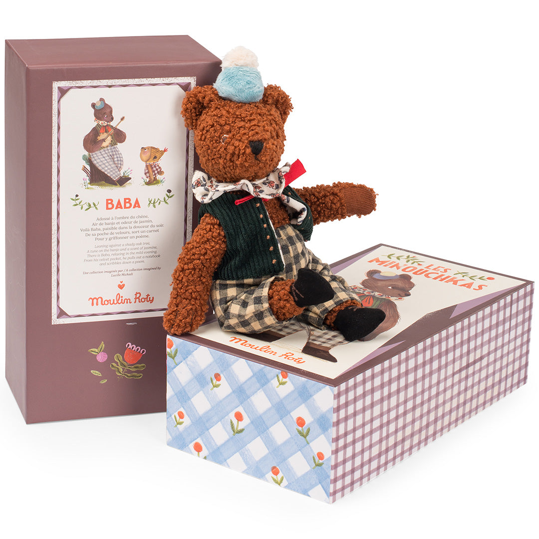 Moulin Roty knuffelbeer Baba in geruite broek en vest, zittend op de open productverpakking.