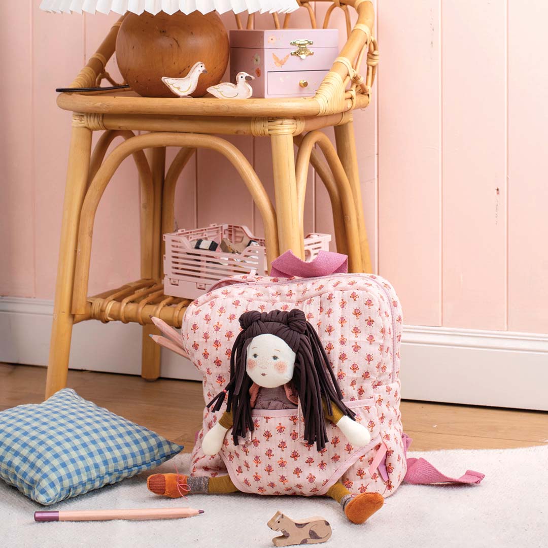 Moulin Roty roze gebloemde poppendraagzak met een pop erin, staand op een vloerkleed in een kinderkamer.