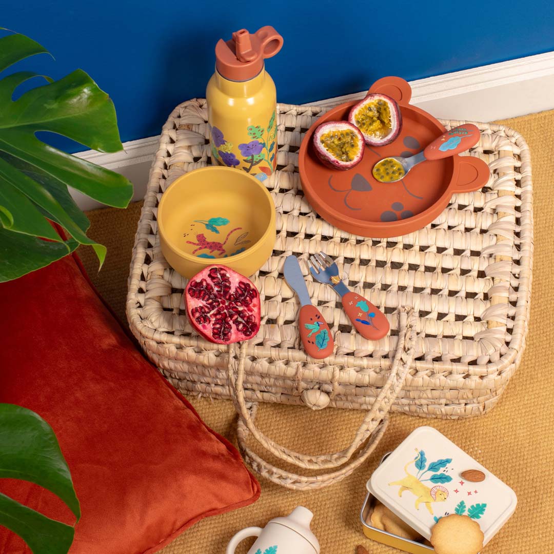 Een collectie van de siliconen serviesset met tijgerbord, kom, beker, waterfles, bestek en lunchbox op een rieten mat.