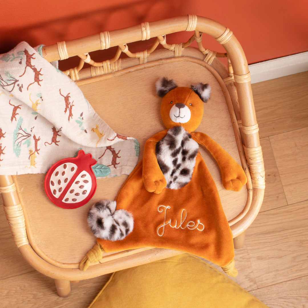 De granaatappel bijtring ligt op een rotan dienblad naast een oranje knuffeldoekje en een doek met dierenprint.