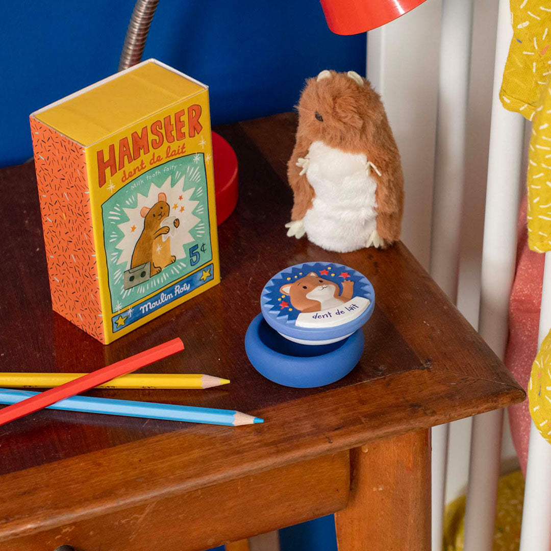 Moulin Roty pluchen hamster tandendoosje met verpakking en een blauw rond tandendoosje op een houten tafel.
