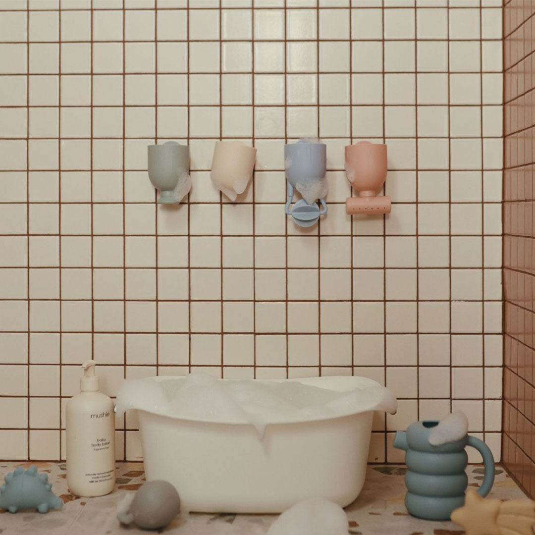 Vier mushie badspeeltjes pour and play hangen aan een tegelmuur boven een babybadje.