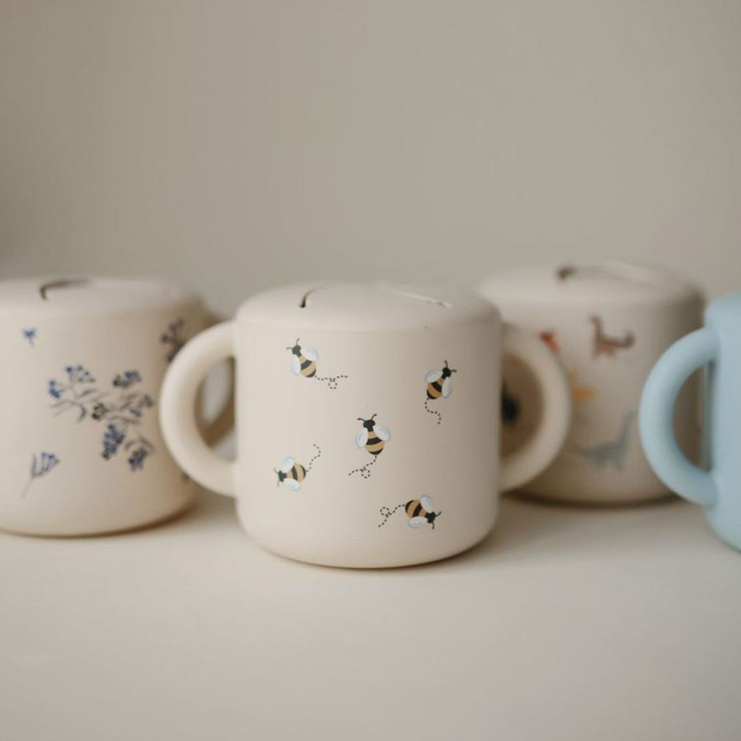 Drie mushie siliconen bekers voor baby's op een rij, waaronder een beige snackbeker met bijenpatroon.