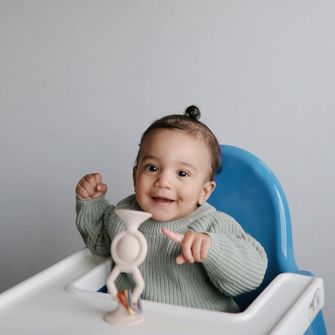 Een lachende baby in een blauwe kinderstoel met het Mushie activiteitenspeeltje op de tray.