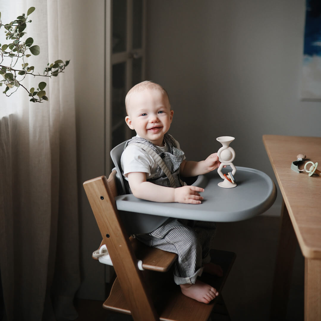 Een lachende baby in een houten kinderstoel die speelt met het Mushie activiteitenspeeltje.