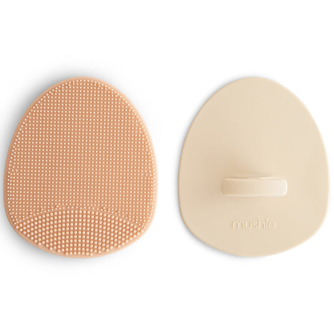 Twee mushie babyborstels tegen berg, één roze met siliconen borstelharen en één beige met gladde achterkant.