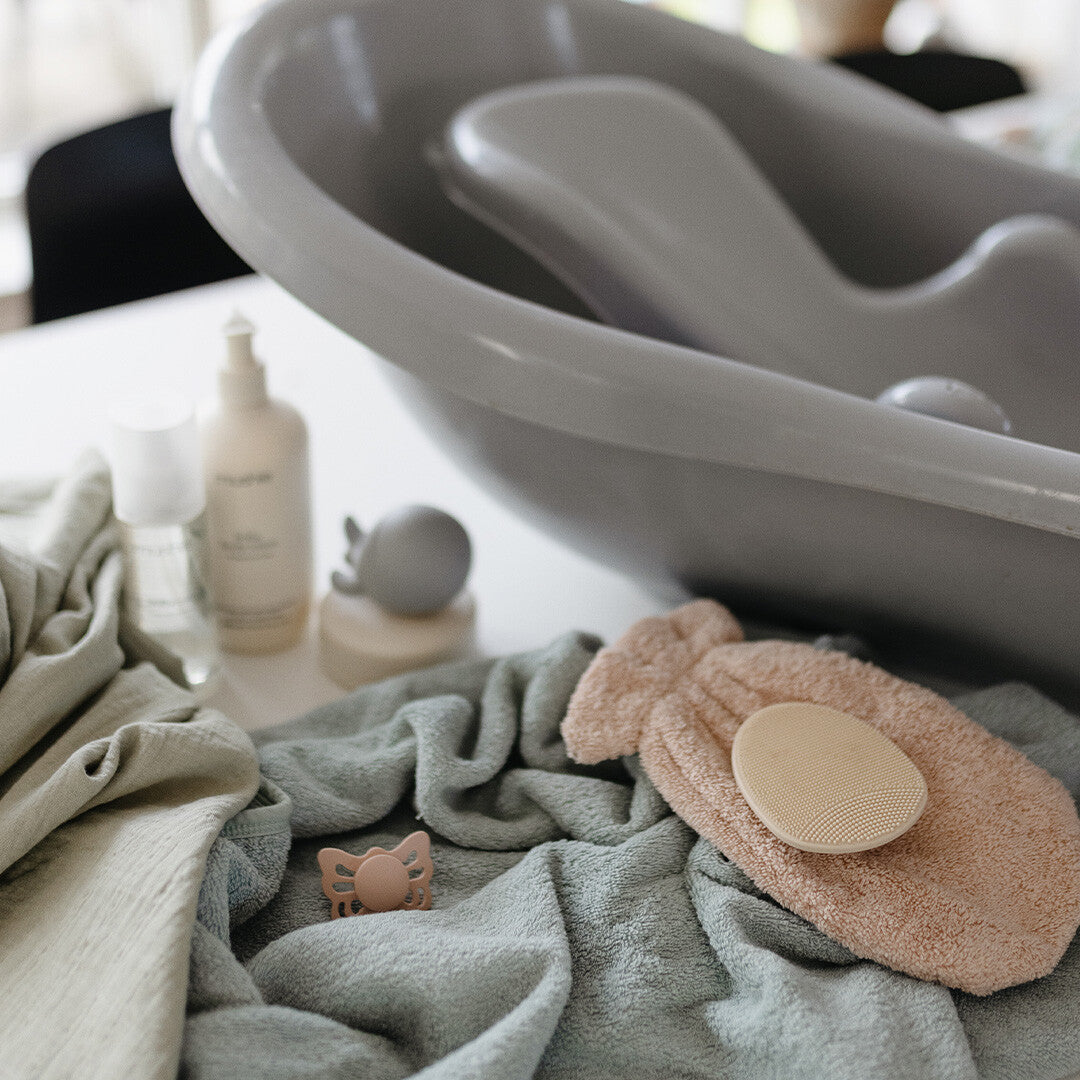 Een collectie baby badartikelen met een grijs babybadje, handdoeken, verzorgingsproducten en een roze mushie babyborstel.
