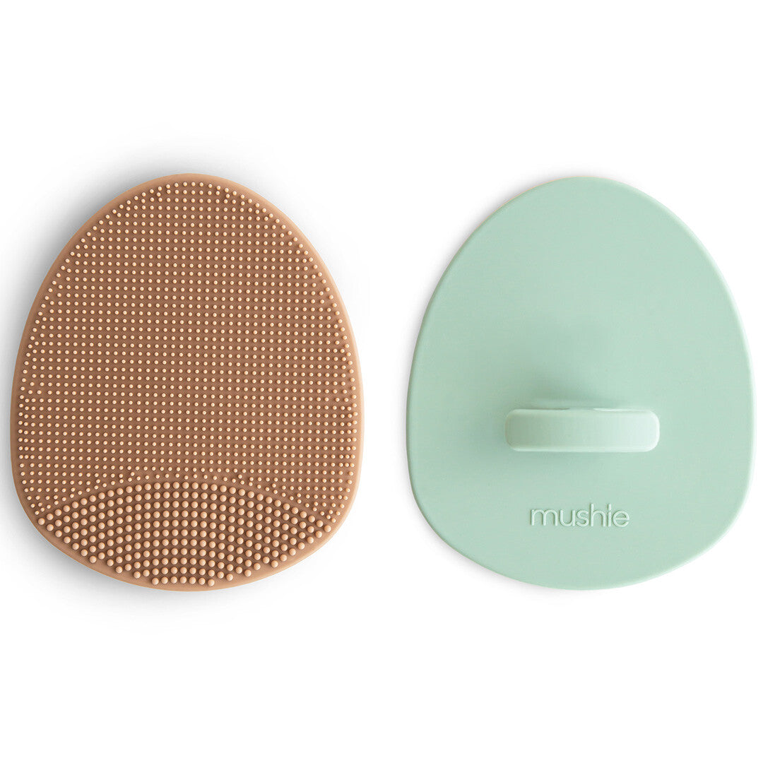 Twee mushie siliconen babyborstels tegen berg, één beige met textuur en één mintgroen met handvat.