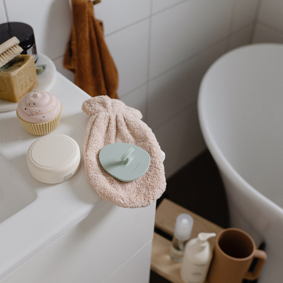 De mintgroene mushie siliconen babyborstel ligt op een roze washandje naast een badkuip.