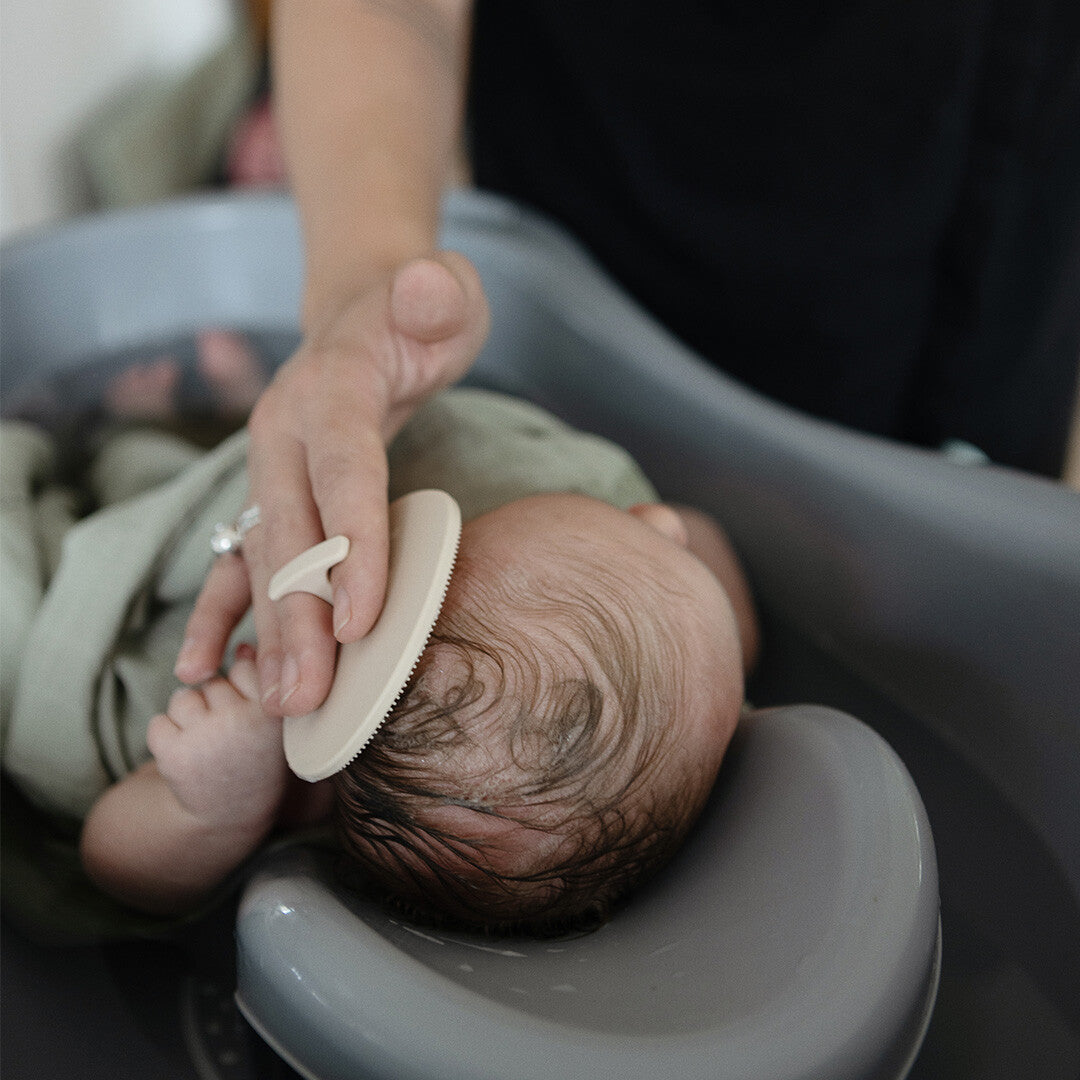 Een volwassene borstelt voorzichtig het hoofdje van een baby met een beige mushie siliconen babyborstel.