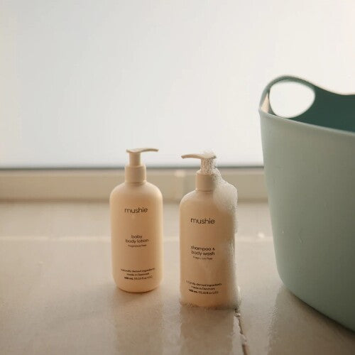 Twee Mushie babyverzorgingsflessen, waaronder babyshampoo, op een natte ondergrond naast een badmand.