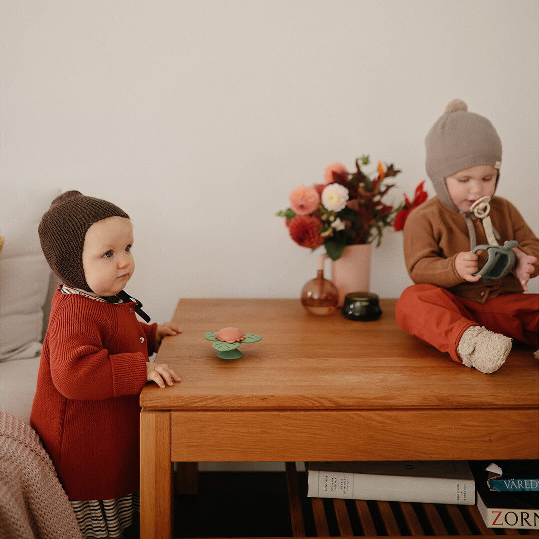 Twee baby's in een kamer, waarvan één bij een houten tafel staat met de mushie babyspeelgoed spinner erop.