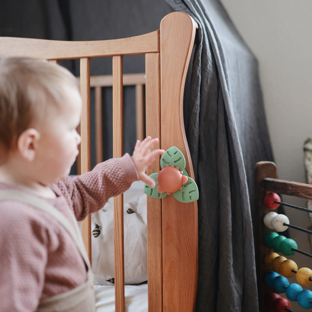 Een baby reikt naar de mushie babyspeelgoed spinner met zuignap die aan een houten ledikant is bevestigd.
